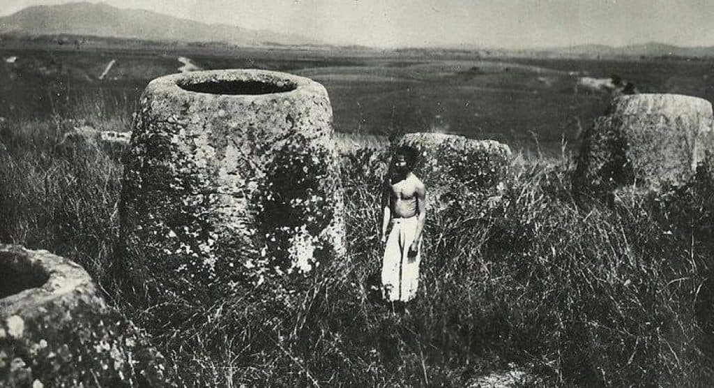 Стара снимка на местен мъж, застанал до гигантски каменен буркан. 